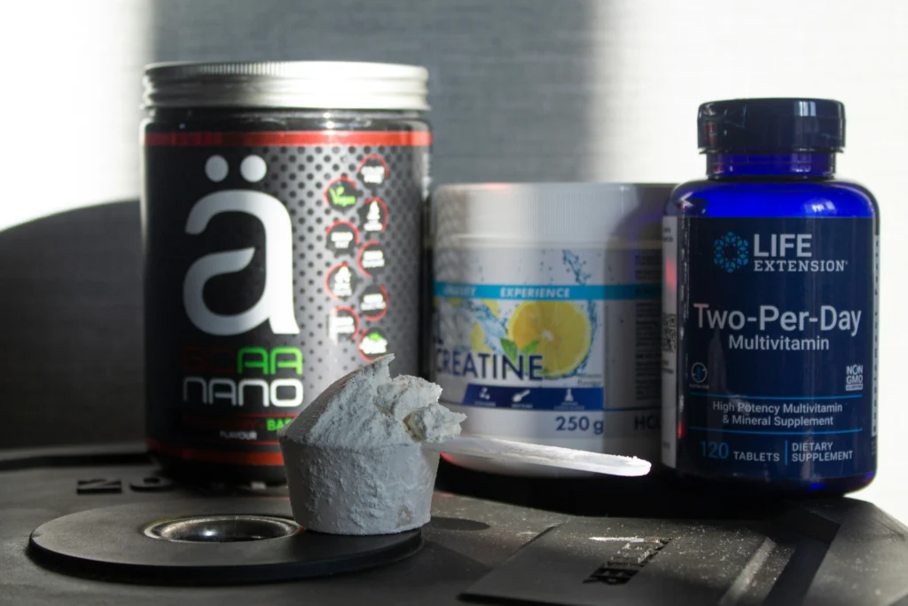 Protein supplements and vitamins on gym scale, including creatine powder, multivitamin tablets, and pre-workout supplements for muscle growth, strength, and daily fitness nutrition